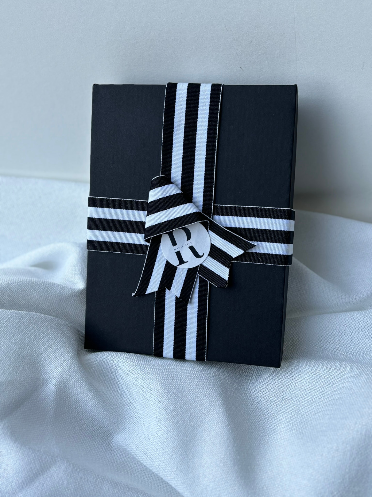 Caja negra mate RMAGIC SILK atada con cinta a rayas blanco y negro y sello redondo con la R